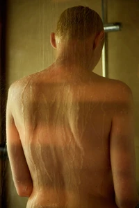 Rear view of a person with wet skin and short hair standing under shower water in a moody interior