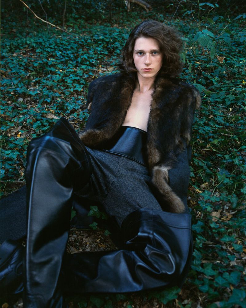 Portrait of a young adult with long brown hair seated outdoors wearing a fur coat, dark top, gray pants and black leather boots in natural lighting.
