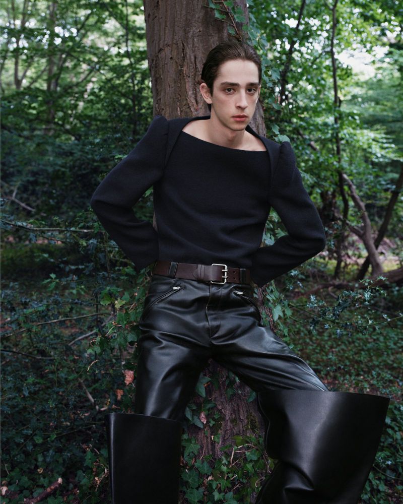 Man posing in a forest wearing a black, long-sleeve, square-neck top, dark leather pants, black boots, and a brown belt with silver buckle.