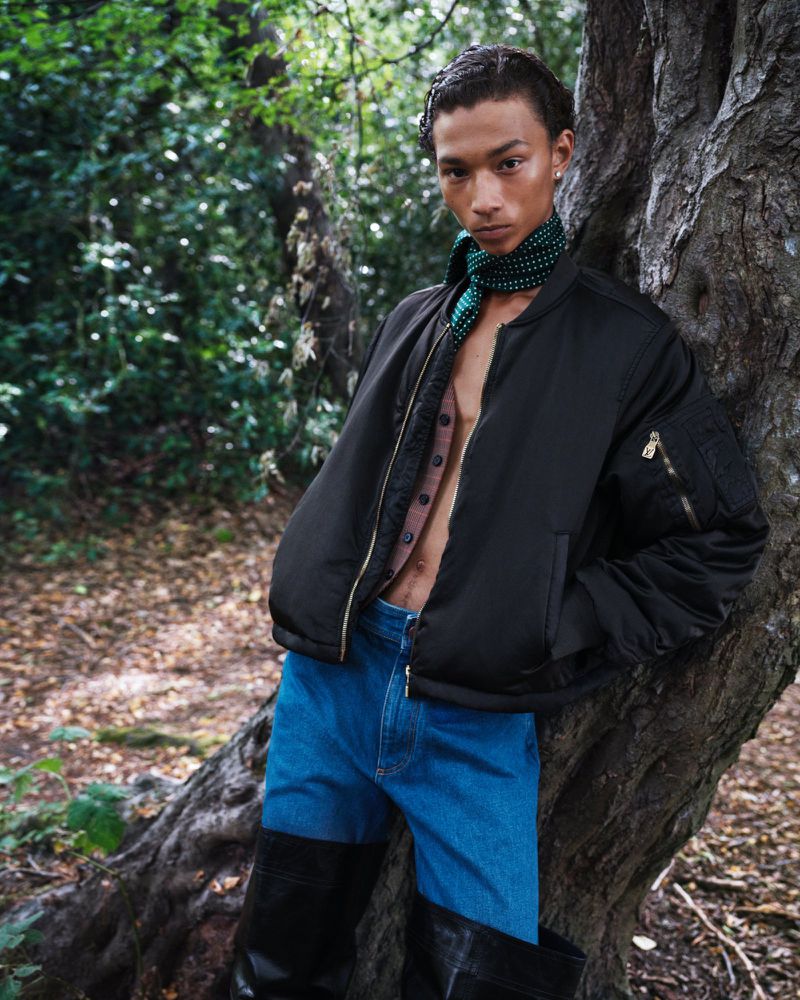 A slender male model leans against a tree in an outdoor forest setting, wearing a black bomber jacket, buttoned brown shirt, and blue jeans.