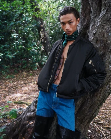 A slender male model leans against a tree in an outdoor forest setting, wearing a black bomber jacket, buttoned brown shirt, and blue jeans.