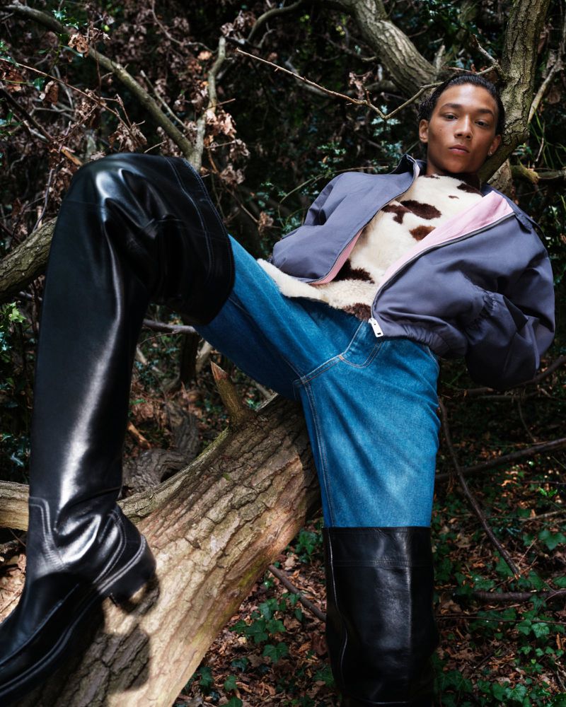 Fashionable young adult in a grey jacket, animal-print top, jeans, and tall black boots, posing on a fallen tree in a forest setting.