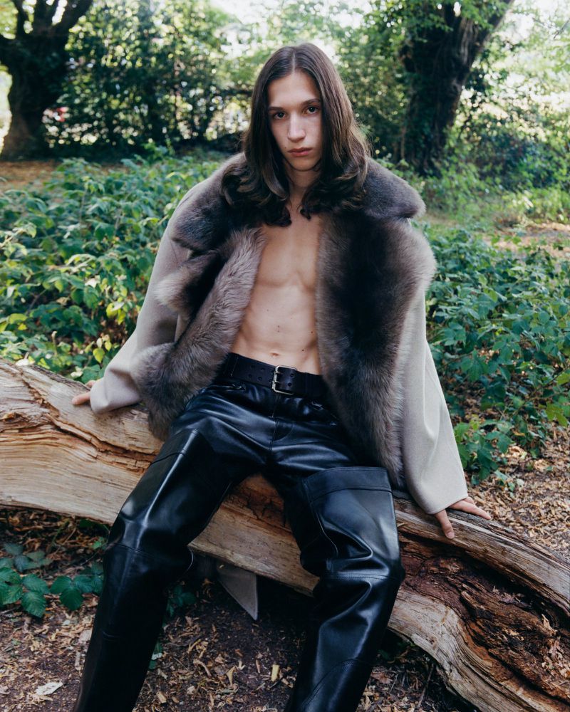 Male fashion model with long hair poses outdoors on a fallen log wearing an open fur-trimmed coat and shiny black pants in a lush forest.