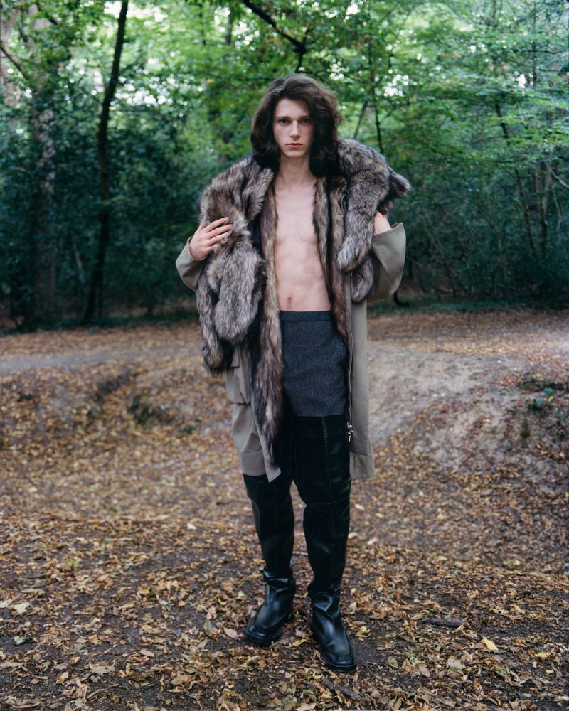 Young adult male posing in a forest wearing a fur stole, coat, black pants and boots, with green trees and brown leaves in background.