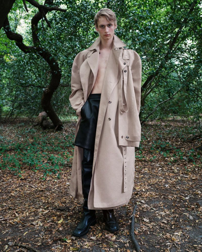 Young adult poses outdoors wearing a beige trench coat, black leather shorts, and black boots in a wooded environment.
