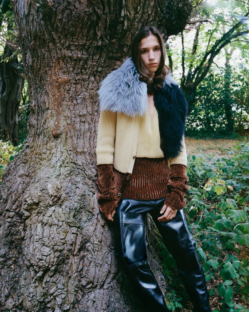 An adult fashion model leans against a tree in a forest setting, wearing a brown and beige color-blocked sweater, faux fur collar, and black pants.