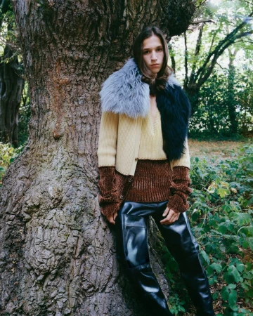 An adult fashion model leans against a tree in a forest setting, wearing a brown and beige color-blocked sweater, faux fur collar, and black pants.