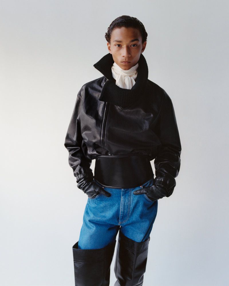 Portrait of a slender person in a black leather jacket, jeans, black gloves, and high black leather boots against a white backdrop.