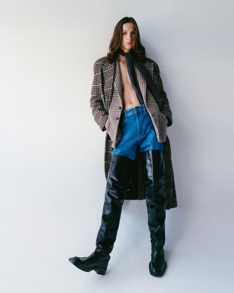 Adult fashion model with long hair poses indoors wearing a patterned coat, blue jean shorts, black scarf, and thigh-high leather boots.