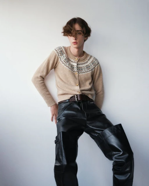 Young adult posing in a tan cardigan with fair isle details, dark leather pants, a belt, and dark curly hair against a white wall.