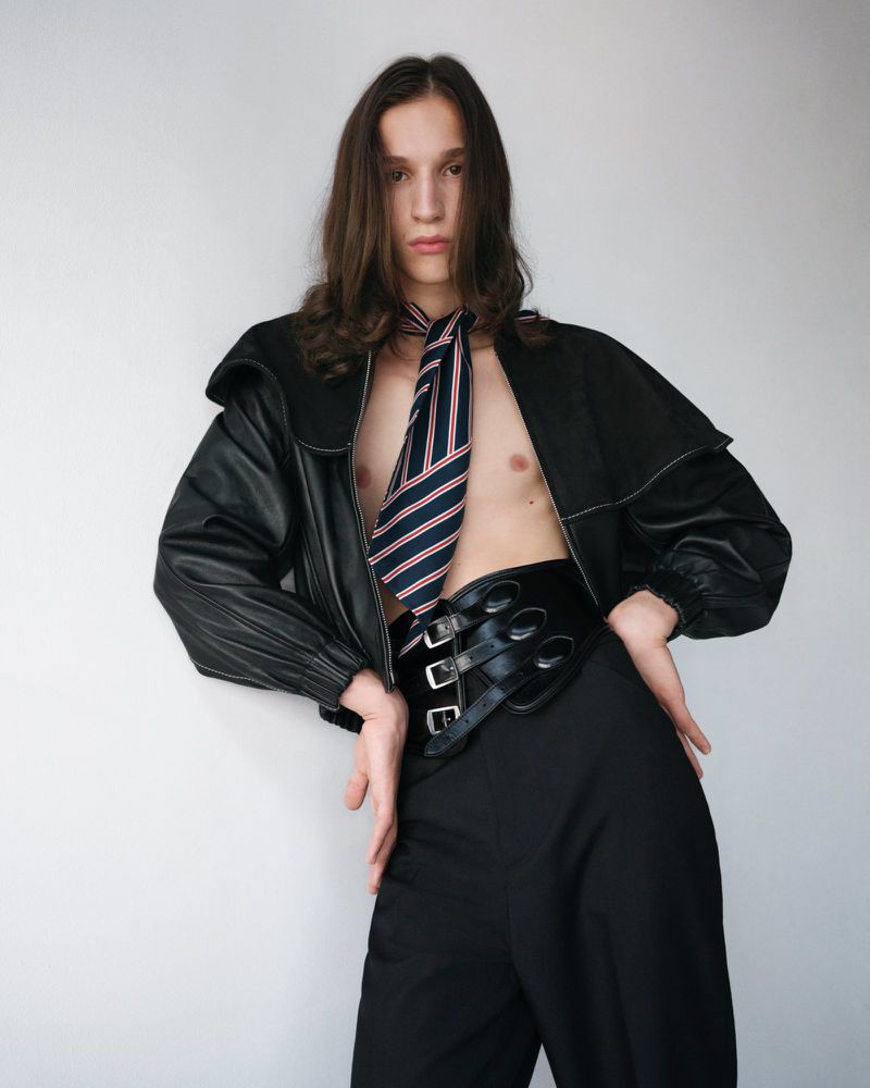 Portrait of a long-haired adult male wearing a black jacket, black pants, and a striped tie indoors against a plain wall.