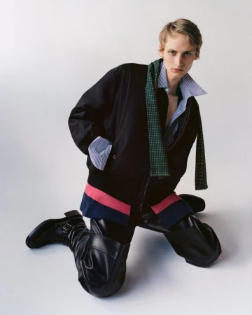Fashionable adult male kneeling in black bomber jacket, striped shirt, dotted scarf, black leather pants and boots against white background.