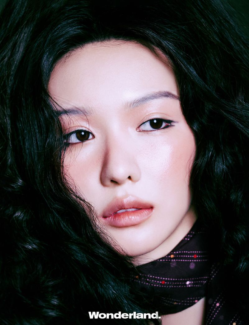 Close-up portrait of a young adult woman with dark, wavy hair, wearing a patterned scarf and soft makeup in a studio setting.