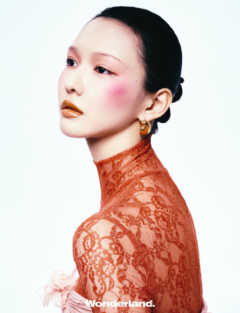 Portrait of a young Asian woman with brown hair, pale skin, and pink blush, wearing a high-necked lace top against a white backdrop.