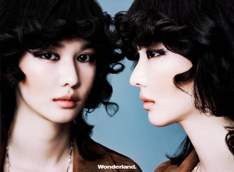 Close-up portrait of two young women with dark, curly hair and dramatic eye makeup against a light blue background. Fashion shot.