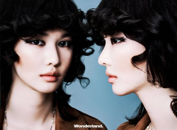 Close-up portrait of two young women with dark, curly hair and dramatic eye makeup against a light blue background. Fashion shot.