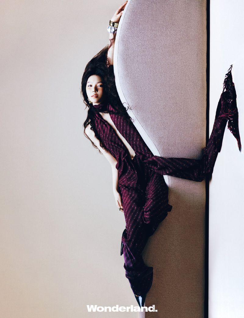 A person in a burgundy outfit leans against a light-colored, padded wall, showcasing a modern, studio fashion portrait with long dark hair.