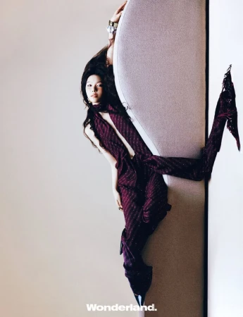A person in a burgundy outfit leans against a light-colored, padded wall, showcasing a modern, studio fashion portrait with long dark hair.