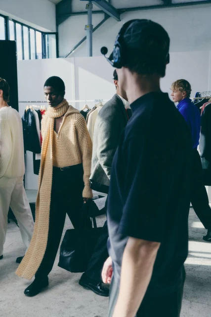 Fashion shoot: several dark and light-skinned male models in knit sweaters and hats backstage, bathed in soft window light.