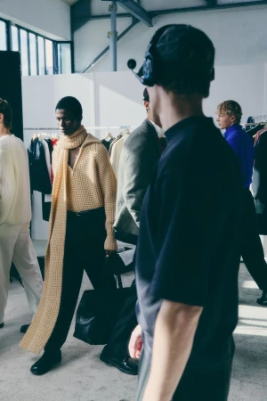Fashion shoot: several dark and light-skinned male models in knit sweaters and hats backstage, bathed in soft window light.
