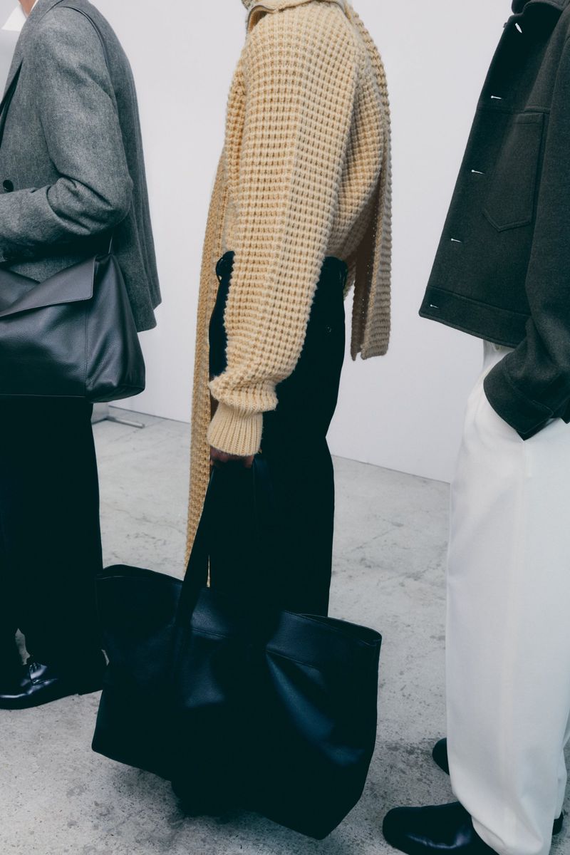 Three people stand indoors wearing black, beige, white clothing; a black leather tote bag is carried by one of the people.