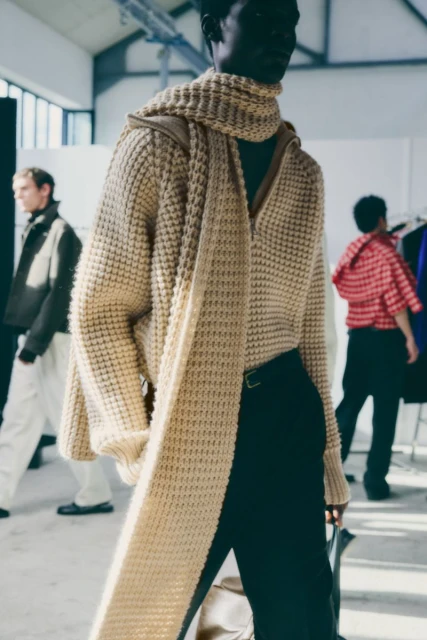 Runway show with a Black male model wearing a beige knit hooded sweater with a long scarf and black pants in a warehouse.