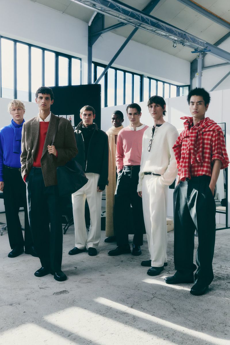Group of male fashion models wearing various stylish outfits stand in a row inside a large industrial building with bright sunlight.
