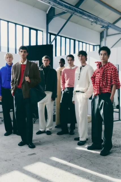 Group of male fashion models wearing various stylish outfits stand in a row inside a large industrial building with bright sunlight.