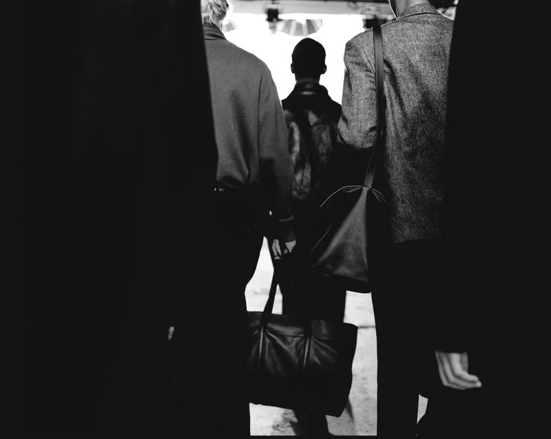 Black and white photo of three people walking away, carrying bags, with an anonymous silhouetted figure in the background.