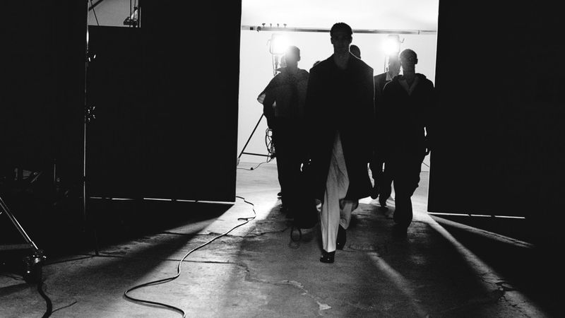 Black and white fashion show scene with a group of models walking towards the camera through a doorway with studio lights in the background.
