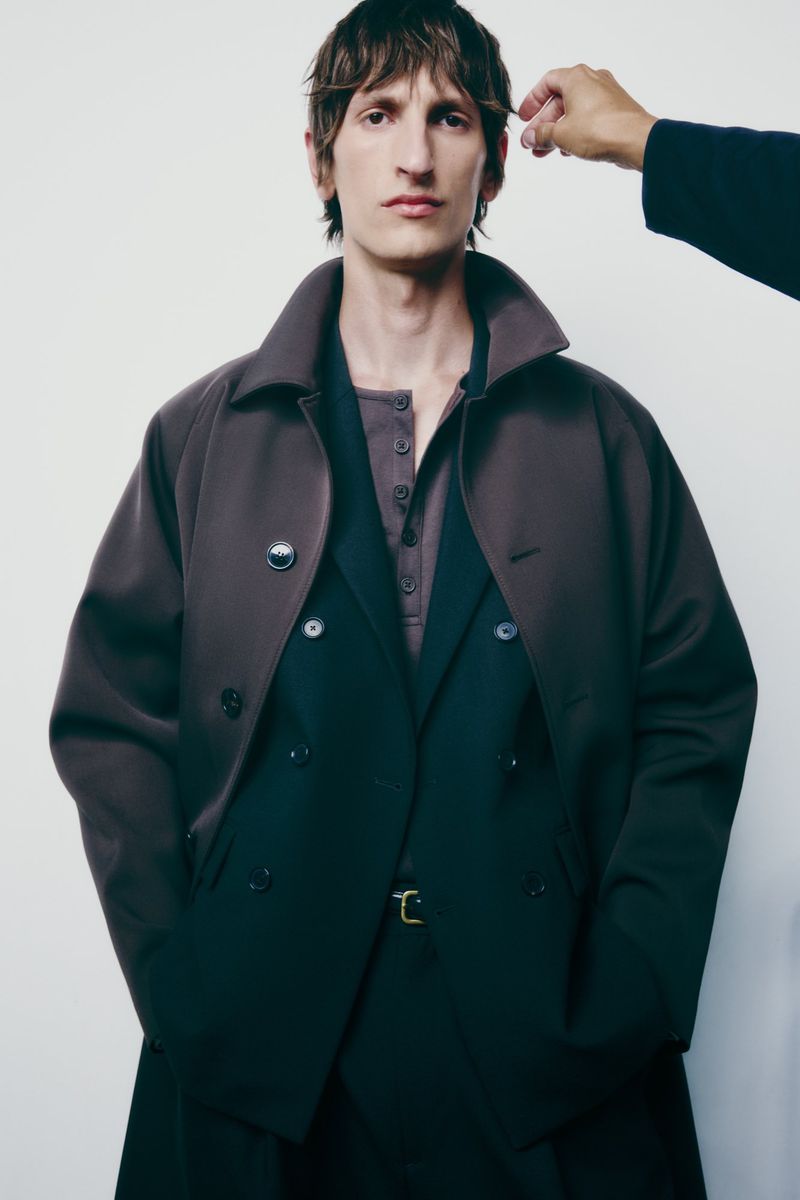 A male fashion model poses indoors wearing a double-breasted coat, suit, and button-down shirt against a neutral backdrop with an adjusting hand.