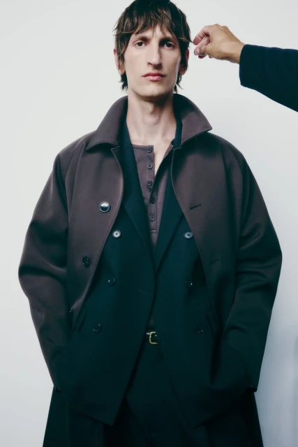 A male fashion model poses indoors wearing a double-breasted coat, suit, and button-down shirt against a neutral backdrop with an adjusting hand.