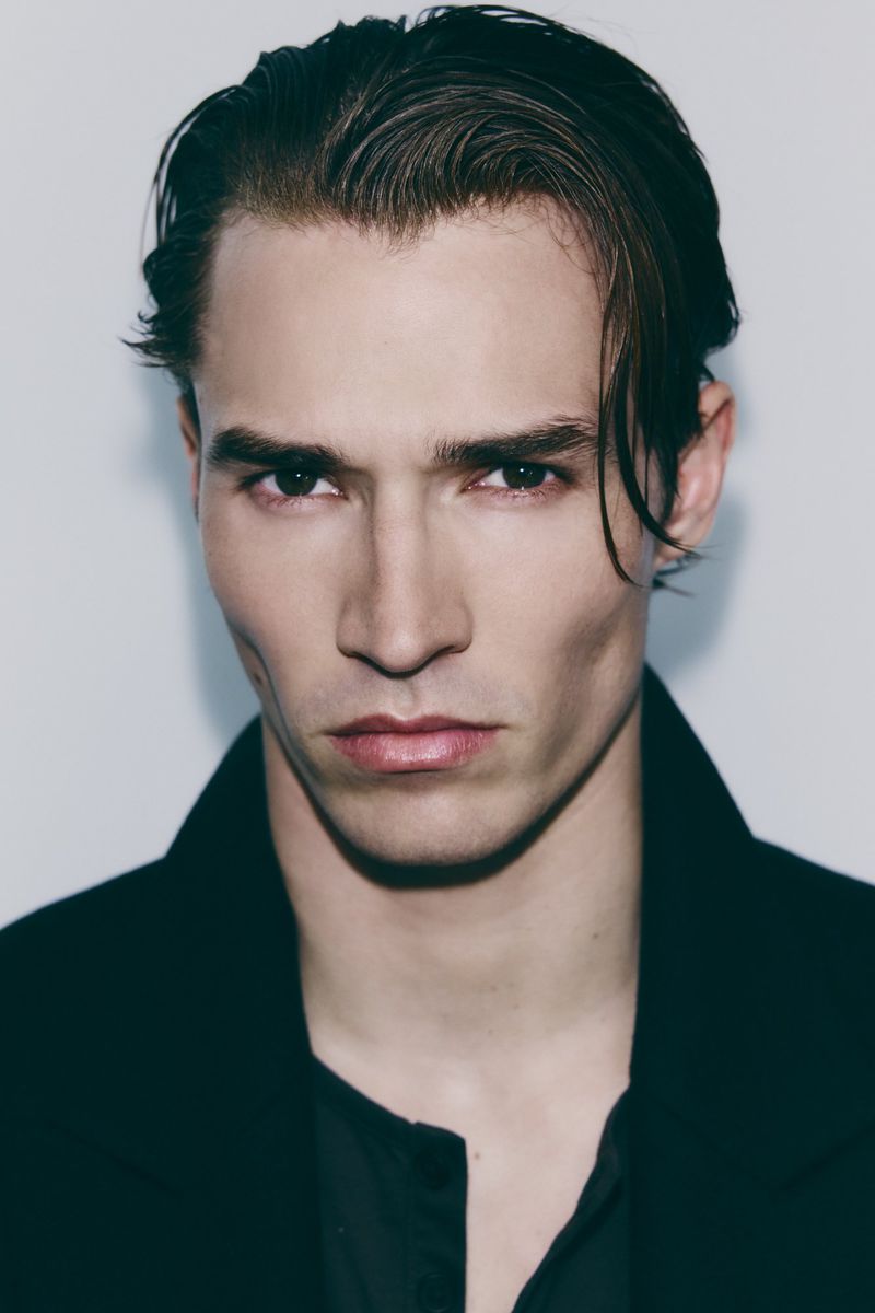 Close-up portrait of a handsome adult male with dark hair wearing a black coat and shirt against a neutral background.