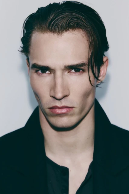 Close-up portrait of a handsome adult male with dark hair wearing a black coat and shirt against a neutral background.