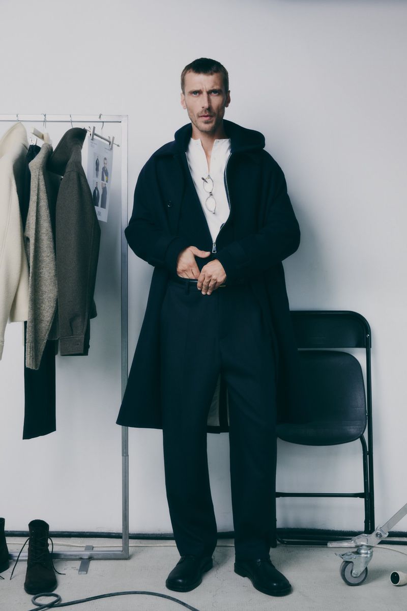 A man in a black coat and pants stands in a photo studio with clothes on a rack and a chair in the background, neutral backdrop.