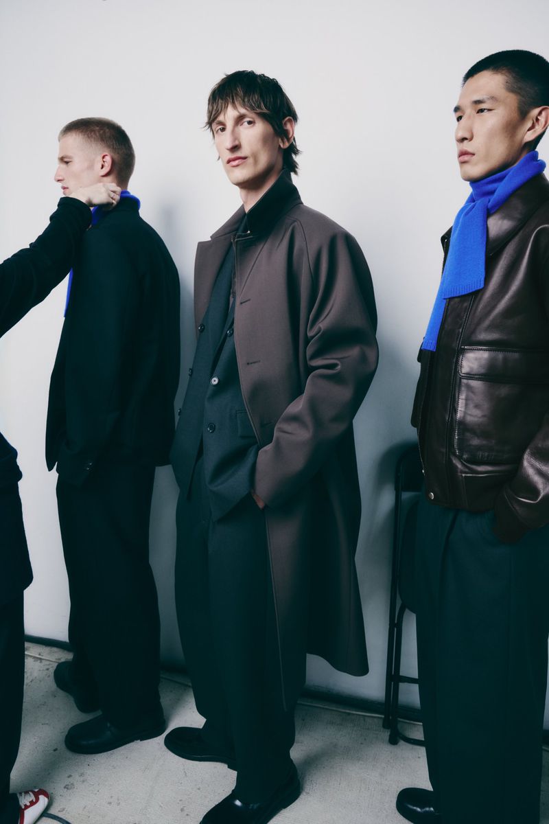 Three adult males are standing indoors wearing fashionable coats and royal blue scarves, with a white wall in the background.