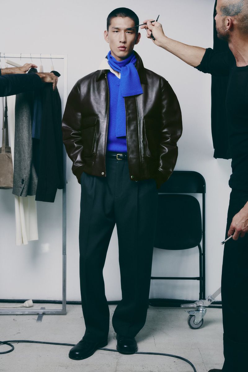 A fashionable Asian male model wears a brown leather jacket and blue scarf in a studio setting with stylists adjusting his hair.