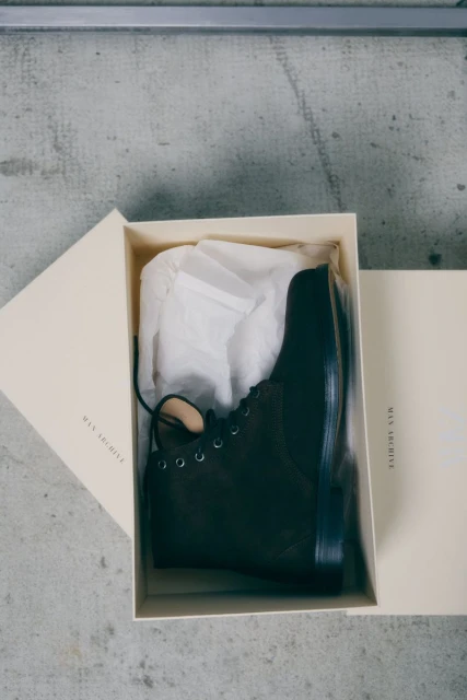 Boxed dark brown leather boots sit on a grey surface with packing paper visible inside, and text visible on both boxes.