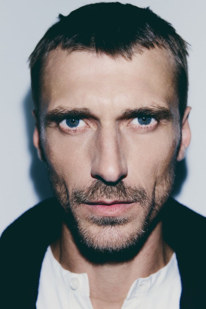 Close-up portrait of a fair-skinned adult male with blue eyes, light brown hair, and a stubble beard against a neutral background.
