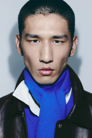 Close-up portrait of an East Asian male model wearing a blue scarf, white collar, and dark jacket against a light background.