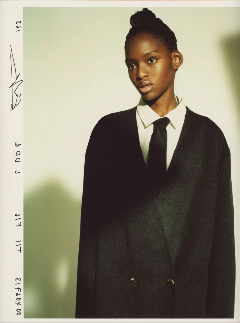 Portrait of a young Black woman in dark jacket, white collared shirt, and tie, against a light gradient background in a studio.