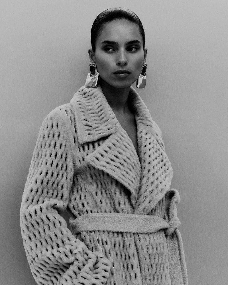 Monochromatic portrait of a female model wearing a textured coat with statement earrings against a light grey background in a studio.