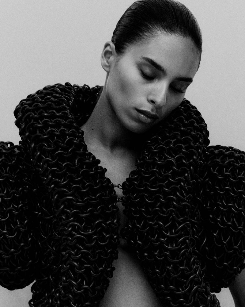 Monochrome studio portrait of a woman with slicked-back hair wearing a chainmail-like jacket with voluminous collar and shoulder elements.