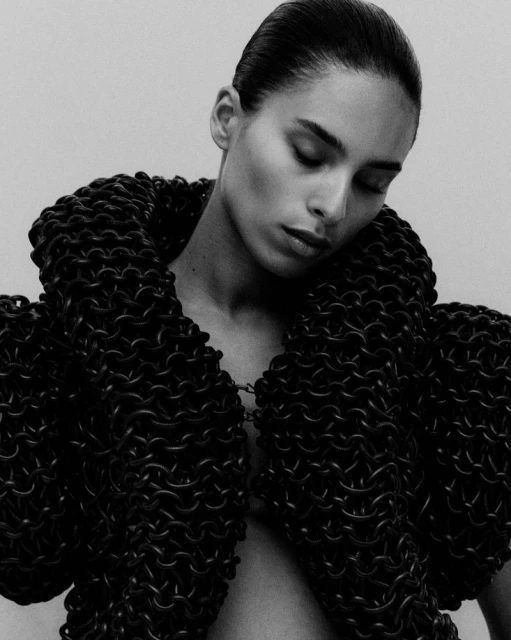 Monochrome studio portrait of a woman with slicked-back hair wearing a chainmail-like jacket with voluminous collar and shoulder elements.