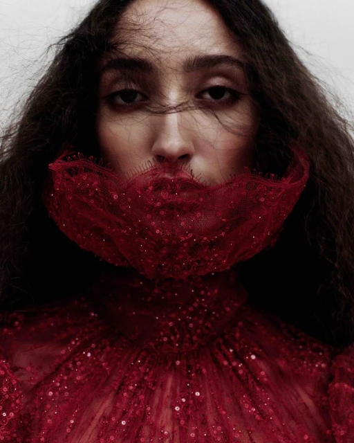 Close-up studio portrait of a woman with dark curly hair wearing a sparkly, reddish-brown, frilled neckpiece and a neutral expression.
