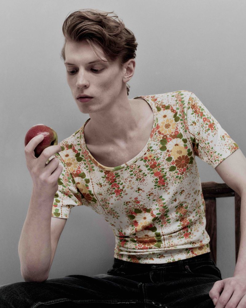 Young man with tousled hair sits indoors holding a red apple, wearing a floral pattern top and dark jeans against a plain background.