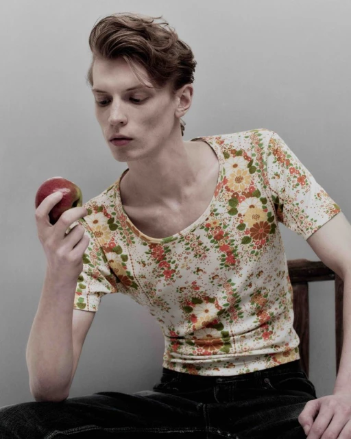 Young man with tousled hair sits indoors holding a red apple, wearing a floral pattern top and dark jeans against a plain background.