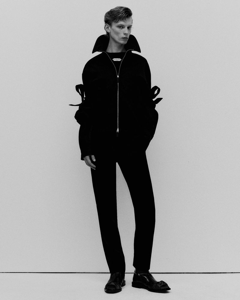 Monochrome portrait of a male model wearing a black zipped jacket with ribbon-tied sleeves against a plain light studio background