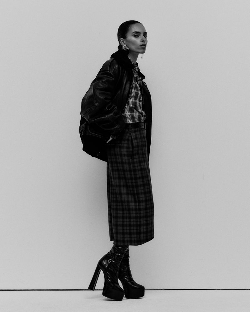 A woman poses in a plaid shirt and skirt, leather jacket, and platform boots against a plain backdrop in a fashion-style studio shot.