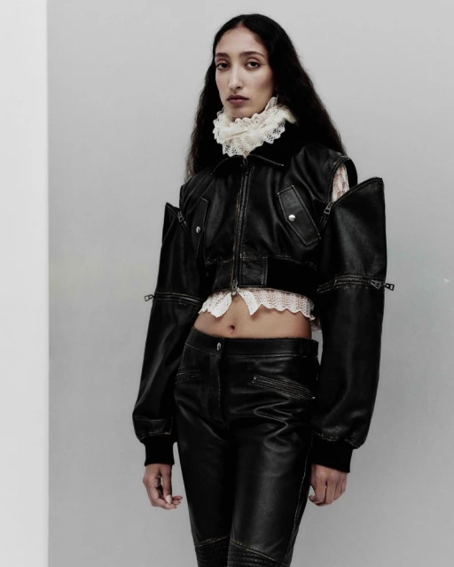 A woman in a black leather jacket and pants poses in a studio with a lace collar, creating a high fashion editorial shot.